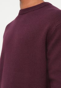 Μπορντό πλεκτό πουλόβερ με στρογγυλή λαιμόκοψη, υφασμάτινη υφή και ραμμένους γύρους. Επικέντρωση στην περιοχή του ώμου και του άνω θώρακα.