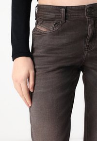 Ciemnobrązowe jeansy z denimu o wąskim kroju, z pięcioma kieszeniami i subtelnym blaknięciem. Charakterystycznym detalem jest czerwone logo na przedniej kieszeni.