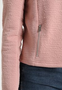 Veste rose texturée avec un col haut, dotée d'une fermeture éclair latérale et de manches longues. Le tissu semble doux et légèrement en relief.