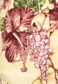 Κίτρινο ύφασμα με μοτίβο από μωβ λουλούδια, που περιλαμβάνει ιβίσκο και ομάδες μικρών λουλουδιών, με καλυμμένα κουμπιά κατά μήκος μιας ραφής.