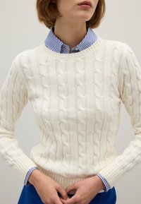 Person in einem weißen Kabelstrickpullover über einem blau-weißen gestreiften Hemd mit Kragen, die Hände am Bauch gefaltet, neutraler Hintergrund.