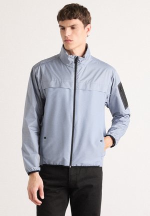 Light blue zip-up jacket with a high collar, elastic cuffs, and a black patch on the left sleeve. Smooth texture and casual fit.