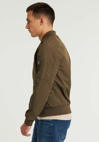 Giacca bomber verde oliva con vita a costine, tasche con zip sulle maniche e texture liscia, indossata sopra una camicia beige e jeans blu.