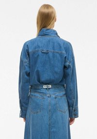 Denim jakna z dolgimi rokavi, svetlo modre barve. Ima strukturiran ovratnik, zapenjanje na gumbih in detajle na hrbtni strani. Vključuje tudi pas.
