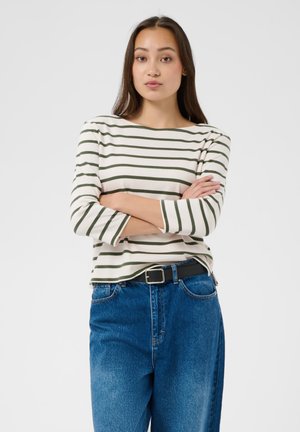 Junge Frau mit langen dunklen Haaren, die ein weiß-grün gestreiftes Oberteil und blaue Jeans trägt, steht mit verschränkten Armen vor einem schlichten Hintergrund.