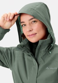 Giacca impermeabile verde con cappuccio, dotata di chiusura con zip, cordoncini regolabili e texture opaca. Logo sul petto.