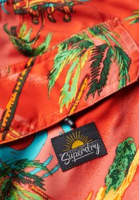 Tessuto dai colori vivaci con un motivo di palme tropicali in tonalità di verde e arancione, con un'etichetta nera che riporta "Superdry" e una grafica del sole.