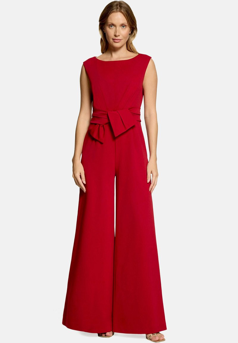 Femme portant une combinaison rouge sans manches à jambes larges avec une ceinture en tissu nouée en nœud à la taille, debout devant un fond uni.