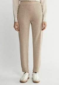 Hellbeige, schmal zulaufende Hose aus einem weichen, strukturierten Stoff. Mit Vorderstaschen und enganliegenden Bündchen, kombiniert mit weißen Sneakers.