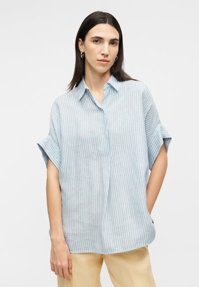 Femme aux cheveux raides et foncés portant des boucles d'oreilles anneaux en or, une chemise ample à manches courtes rayée bleu clair et blanc, et un pantalon beige.