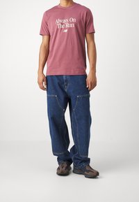 Man met een mauve T-shirt met de tekst "Altijd Op De Ren", losse blauwe denim jeans met frontzakken, en bruin-zwarte sneakers.