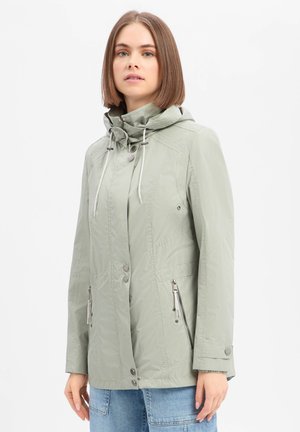 Vrouw met steil bruin haar die een lichtgrijze capuchonjas draagt met ritssluitingen aan de zijzakken en drukknopen, staand tegen een witte achtergrond.