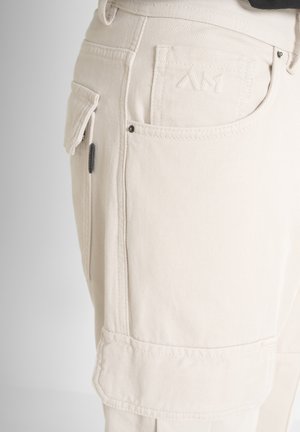 Pantaloni cargo beige in tessuto testurizzato, con tasche laterali e un logo cucito sul retro. Cuciture visibili e dettagli in metallo.