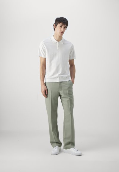 Hvitt kortermet poloskjorte med krage, kombinert med lysegrønne bukser. Mannen står med hendene i lommene, iført hvite joggesko.