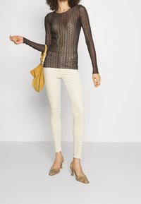 Top in pizzo nero trasparente a maniche lunghe con righe verticali, abbinato a jeans skinny color crema. Gli accessori includono una borsa gialla e tacchi con motivo.