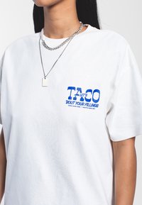 Bílá bavlněná tričko s modrou grafikou, na které je nápis "POJĎME SE BAVIT O TVÝCH POCITECH." Krátké rukávy a pohodlný střih. Viditelné stříbrné náhrdelníky.