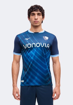 Man wearing a blue sports jersey with light blue stripes, white "Vonovia" text, and a shield logo on the chest, standing with arms by his side.