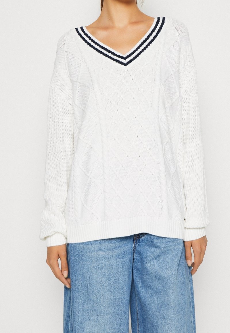 Bílý pletený svetr se siluetou ve tvaru V, s námořnicky modrými a bílými pruhovanými lemy, vzory v podobě kosočtverců a copánků, s dlouhými rukávy.