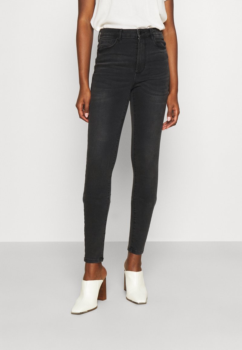 Jeans skinny pretos de cintura alta com uma textura suave, apresentando cinco bolsos e um design clássico de denim, combinados com mules brancos de salto bloco.