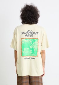 Beige oversized t-skjorte med et grønt grafisk motiv rammet inn i oransje, med teksten "ALL CONDITIONS GEAR" og "Big Bend, TEXAS" nedenfor.