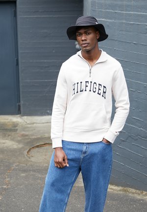 Felpa con zip color crema con la scritta "HILFIGER", indossata sopra un paio di jeans a gamba larga blu chiaro. Un cappello a secchiello nero contrasta con l'outfit.