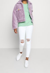 Faux kožešinová fialová bunda, zelené tričko s crew neck, bílé seprané skinny jeans a bílé tenisky, které předvádějí jemné textury a vrstvený styl.