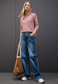 Rosa Strickpullover, blaue weit geschnittene Jeans mit Seitstripes, braune Wildleder-Tasche mit strukturierten Griffen und weiße Sneaker.