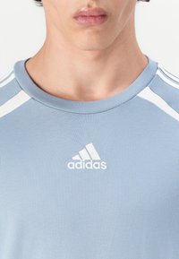 Šviesiai mėlynas sportinis marškinėliai su rausvu apykaklės kraštu, baltais akcentais ant pečių ir išsiuvinėtu baltu Adidas logotipu ant krūtinės.