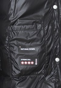 Tessuto della giacca a vento nera con una finitura lucida. Presenta due etichette: "NATURAL DOWN" e "EXTREME CHILL" con cinque icone di foglie d'acero e un grafico del termometro.