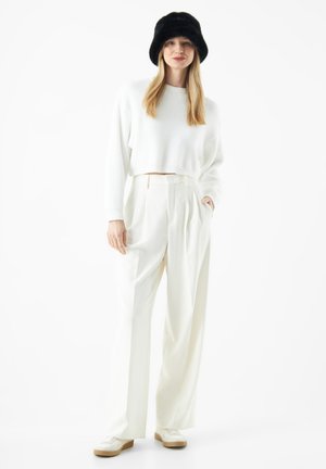 Witte cropped trui, crème wijde broek en crème sneakers. Zwarte bonten bucket hat voegt contrast toe. Glad oppervlak overal.