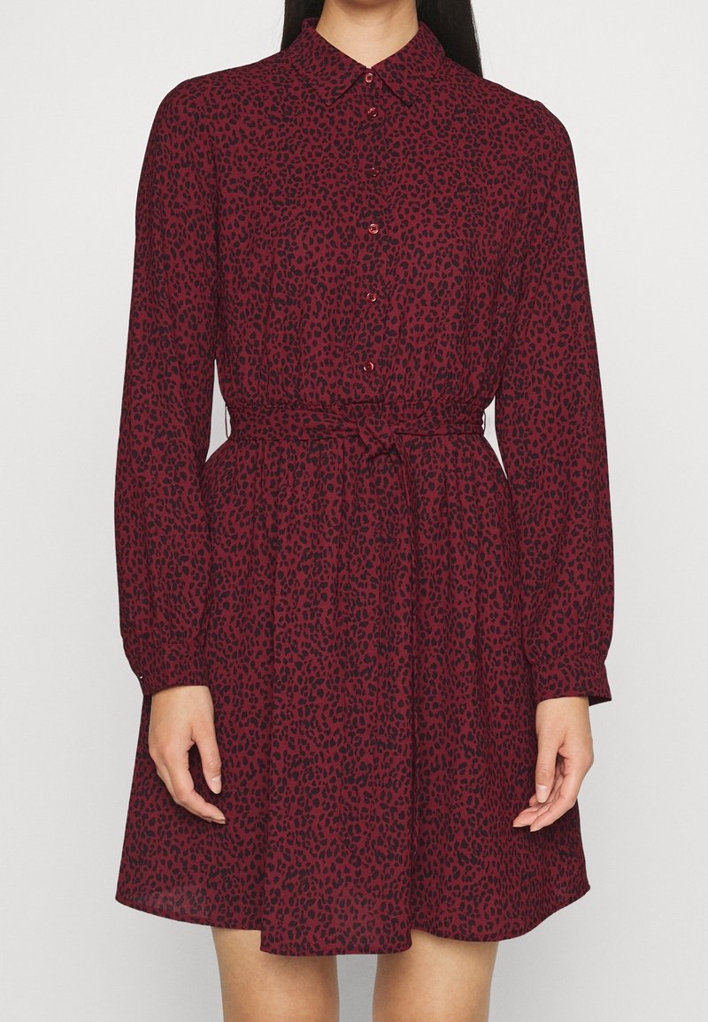 Femme portant une robe bordeaux à manches longues avec imprimé léopard noir, boutonnée devant, col et ceinture nouée à la taille.