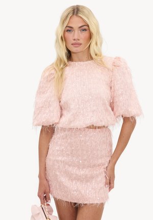 Femme blonde porte un haut rose texturé et duveteux ainsi qu'une jupe assortie, tenant un petit sac à main rose à côté d'elle.
