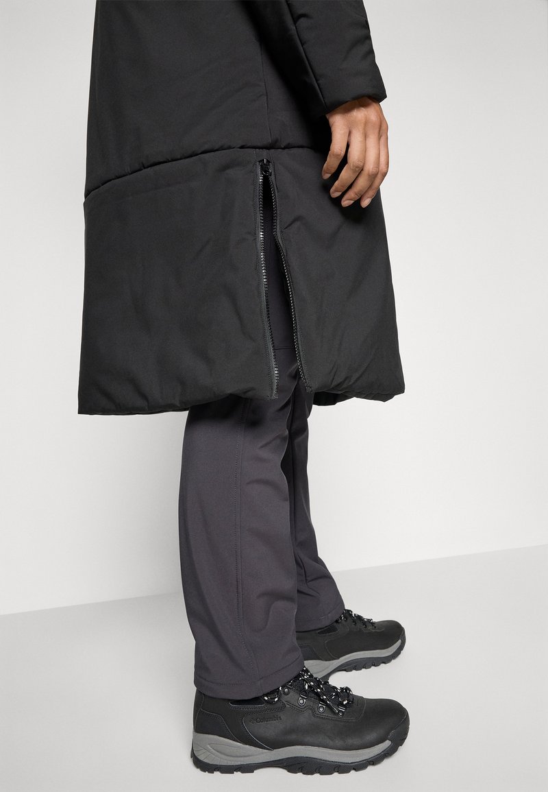 Person mit schwarzen Wanderschuhen und grauen Hosen, die einen langen schwarzen Mantel trägt, der einen teilweise geöffneten Seitenschluss hat.