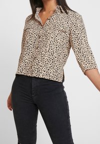 Animal print blouse in beige and black, featuring a button-up front, collar, and two chest pockets; fabric appears smooth and lightweight.