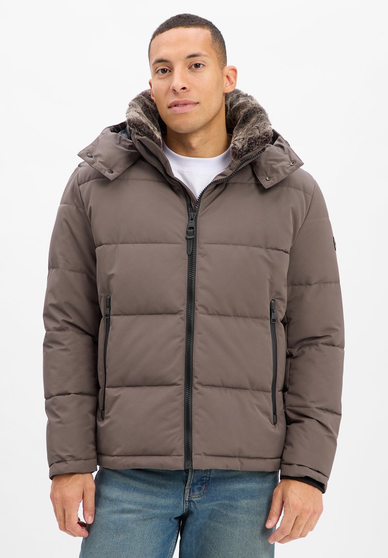Mann in einer grauen gepolsterten Winterjacke mit fellgefütterter Kapuze, Frontreißverschluss, zwei Reißverschlusstaschen und blauen Jeans vor einem weißen Hintergrund.