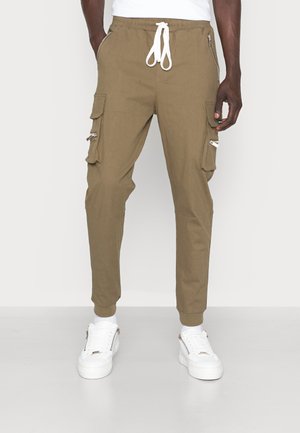 Homme portant un pantalon jogger cargo kaki avec un cordon blanc et des baskets blanches, debout devant un fond clair uni.