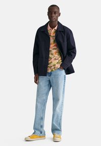 Námořnická bunda s knoflíky, vzorovaná růžová košile s palmami, světle modré džíny a žluté tenisky s bílou podrážkou.