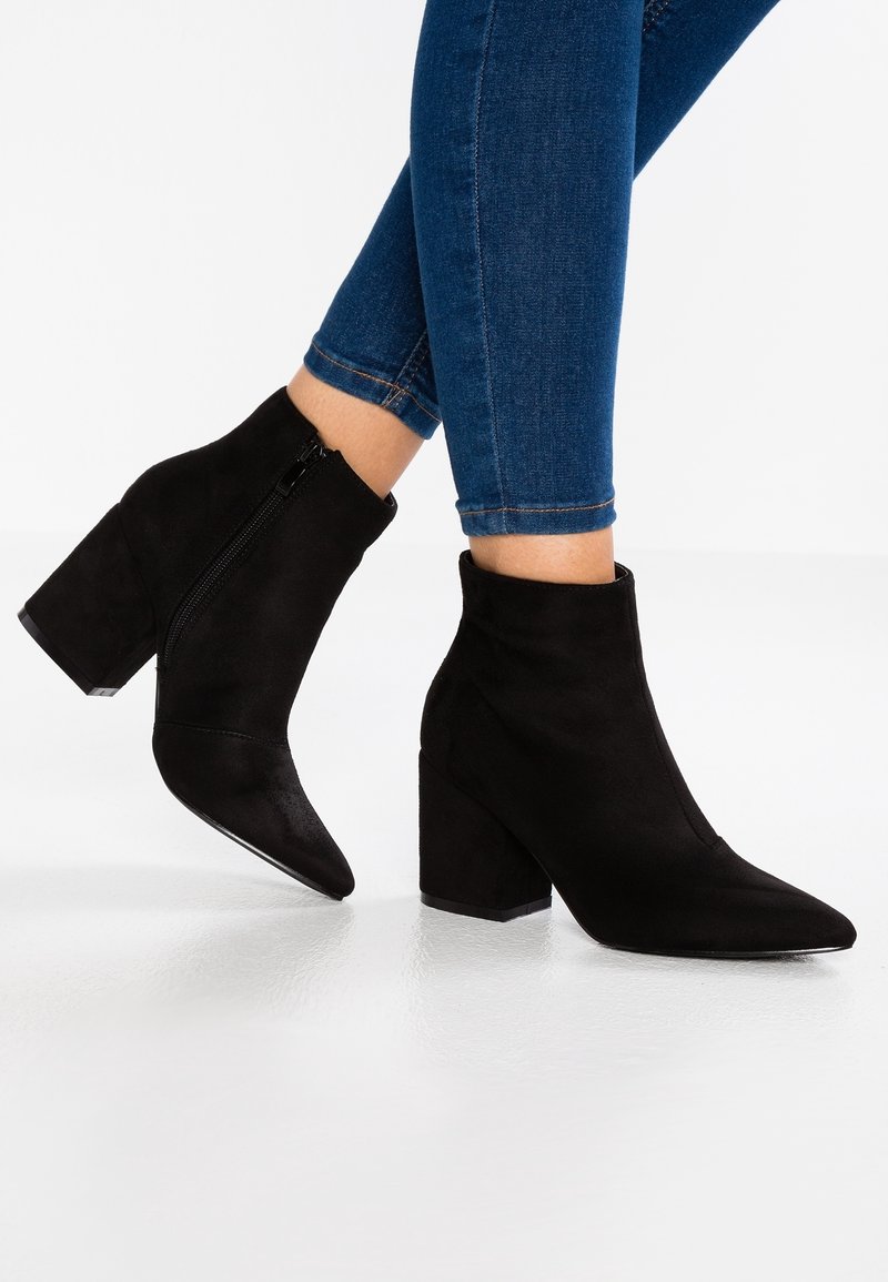 Bottes cheville en daim noir avec bout pointu et talon carré. Dotées d'une fermeture éclair sur le côté et d'une texture lisse, portées avec un jean bleu ajusté.