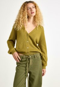 Olivgrön stickad cardigan med V-ringning, knappfram, och ribbade ärmslut, parat med matchande gröna byxor. Texturdetalj synlig.
