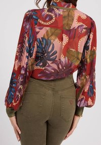 Vrouw in een doorschijnende rode blouse met tropische bladeren en kettingpatronen, gecombineerd met hoog aangesloten olijfgroene broeken met achterzakken.