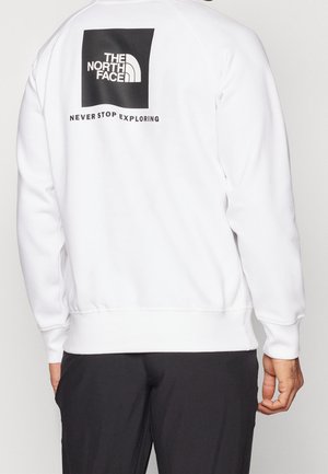 Osoba ubrana w białą bluzę z kapturem z czarnym logo „The North Face” oraz napisem „Never Stop Exploring” na górnej części pleców, w połączeniu z czarnymi spodniami.