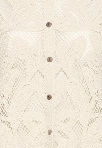 Tissu crochet couleur crème avec motifs floraux et quatre petits boutons marron alignés verticalement au centre.