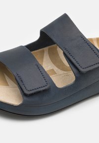 Temno modre usnjene sandale z dvema nastavljivima Velcro trakovama, odprto zasnovo toe in obdanimi bež podplati, ki ima subtilen vzorec.