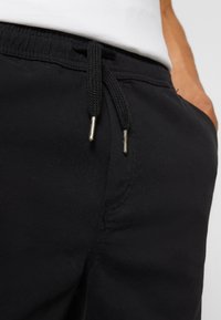 Gros plan sur un pantalon noir à cordon de serrage avec des aglets en métal, porté par une personne ayant une main dans la poche et une chemise blanche rentrée.