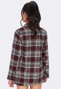 Frau mit dunklem Haar in einem hohen Pferdeschwanz, die große rote Creolen und ein bordeauxfarbenes und weißes Karoblazer-Kleid trägt, mit dem Rücken zur Kamera.