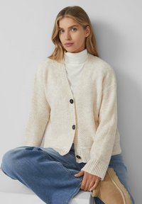Blonde Frau mit blauen Augen, die einen cremefarbigen Strickcardigan, einen weißen Rollkragenpullover, blaue Jeans und beigefarbene Ankle Boots trägt, sitzt vor einem grauen Hintergrund.