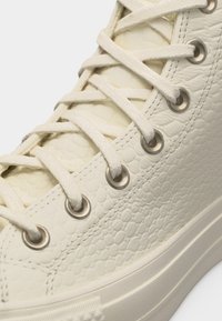 Κοντινό πλάνο κρεμ-χρωματιστού υφασμάτινου ψηλού αθλητικού παπουτσιού με μεταλλικά οπές και χοντρές, επίπεδες κορδόνια σε σταυρωτό μοτίβο.