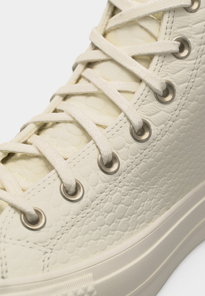 Primo piano di una sneaker alta color crema con trama testurizzata, occhielli metallici e lacci spessi e piatti incrociati a X.