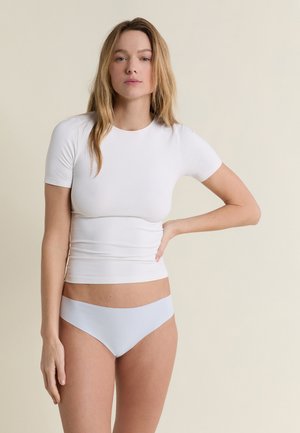 Femme aux longs cheveux blonds, portant un t-shirt blanc ajusté à manches courtes et un sous-vêtement blanc sans coutures, debout devant un fond beige.