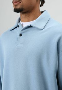 Hellblaues Poloshirt aus weichem Stoff mit einem breiten Kragen, zwei schwarzen Knöpfen und einer dezenten Struktur.